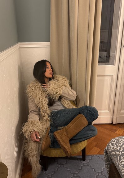 Abrigo beige de piel sintética con cuello ancho, camisa gris, vaqueros azules y botas marrones hasta la rodilla. Sentada en una silla con estampado, cerca de una cortina y una puerta.