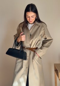 Smėlio spalvos medvilninis tranšėjos paltas su sagomis ir diržu juosmens srityje, derinamas su juoda rankine ir laikomas ruda knyga. Minimalistinis dizainas.