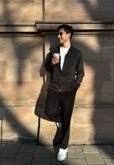 Camisa negra de botones sobre una camiseta blanca, combinada con pantalones negros y zapatillas blancas. Sosteniendo una taza de café, apoyado contra una pared de piedra.