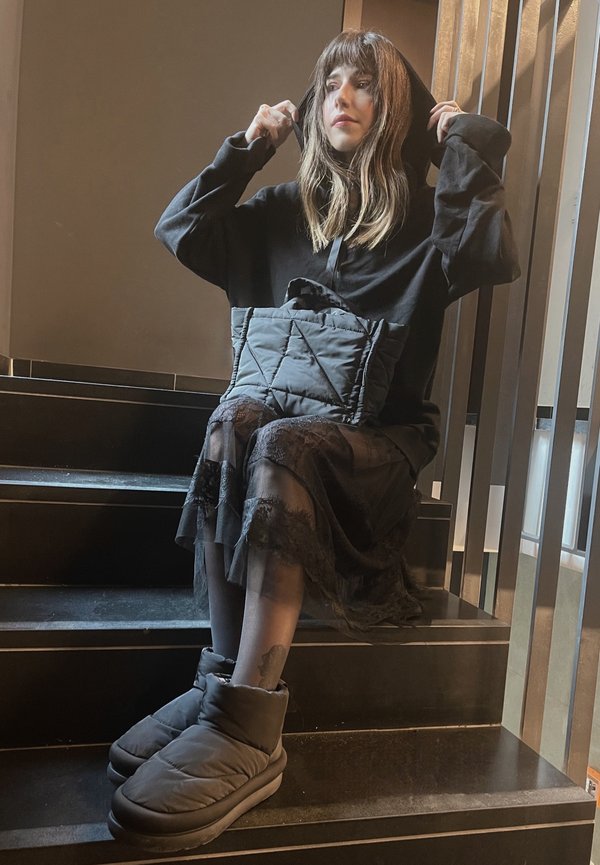Black oversized hoodie with a hood, quilted black bag, layered sheer black skirt with lace trim, and bulky black slippers on stairs.