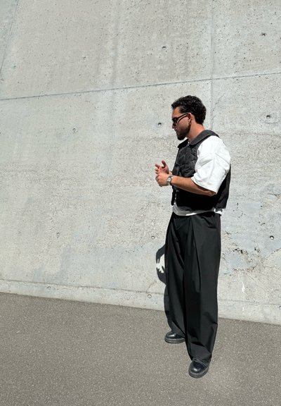Chaleco acolchado negro sobre una camisa blanca de gran tamaño, combinado con pantalones anchos negros y zapatos negros, de pie contra una pared de concreto.
