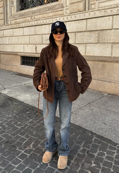 Blazer de ante marrón sobre una camiseta beige, combinado con jeans azul claro y botas beige. Los accesorios incluyen una gorra negra y un bolso marrón.