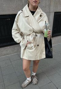Trench-coat beige avec taille nouée et gros boutons, porté sur un haut noir. Associé à des baskets grises et un sac noir. Boisson verte à la main.