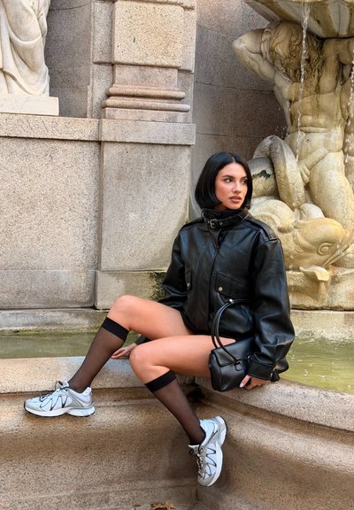 Chaqueta de cuero negra con dos bolsillos delanteros, combinada con calcetas negras transparentes hasta la rodilla y zapatos deportivos plateados. Pequeño bolso de mano negro.
