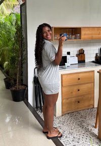 Femme en tenue décontractée grise et tongs souriante, tenant une tasse bleue dans une cuisine avec des armoires en bois et un sol en carrelage à motifs.