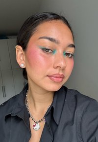 Retrato de una persona con una camisa negra, que lleva un collar de Hello Kitty y pendientes. El maquillaje incluye sombra de ojos en tono azul verdoso y un rubor rosa.
