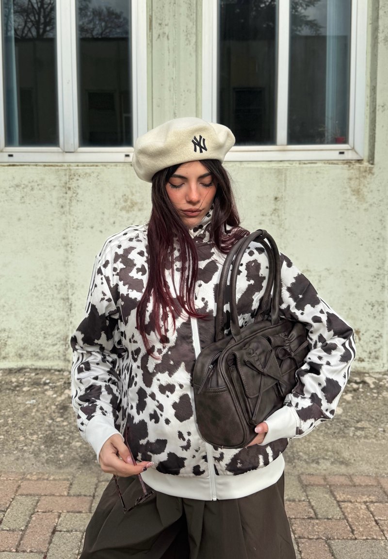 Chaqueta con estampado de vaca en blanco y negro y cremallera blanca, combinada con un bolso de cuero marrón oscuro con múltiples bolsillos. Boina blanca en la cabeza.