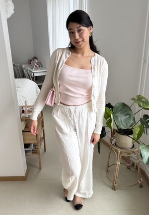 Pantalones de lino blancos ligeros combinados con una camiseta de tirantes rosa de canalé y un cárdigan crema. Accentuados con unas bailarinas negras y beiges y un pequeño bolso rosa.