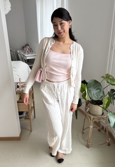 Pantalones de lino blancos ligeros combinados con una camiseta de tirantes rosa de canalé y un cárdigan crema. Accentuados con unas bailarinas negras y beiges y un pequeño bolso rosa.