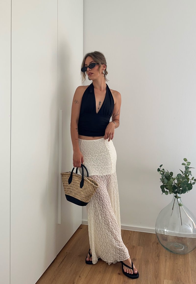 Femme portant un haut noir dos nu et une jupe en dentelle blanche tenant un sac tressé, debout près d'un mur blanc et d'un grand vase en verre avec des branches vertes.