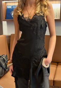 Γυναίκα με μαύρο φόρεμα με δαντέλα και χρυσό σταυρό, κρατώντας μαύρο ύφασμα και μπεζ βραχιόλι, στέκεται μπροστά από έναν καφέ καναπέ.