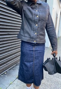 Giacca di jeans nera con bottoni dorati, indossata sopra una gonna di jeans blu scuro. L'outfit presenta un design strutturato e dettagli di cuciture visibili.