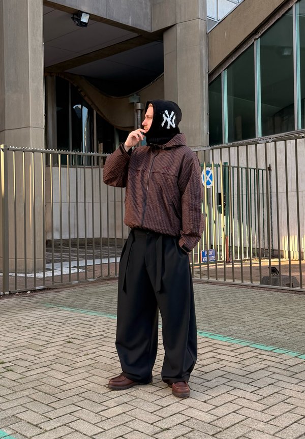 Mees, kes kannab ruudulist pruuni jopet, lahtisi musti pükse, pruune jalanõusid ja musta balaklaavi valge NY logoga, seisab sillutisel tänaval metallpiirde lähedal.