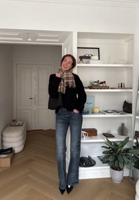 Femme portant une écharpe à carreaux, un pull noir et un jean, debout à l'intérieur près d'une étagère blanche avec des livres, des plantes et des objets décoratifs.