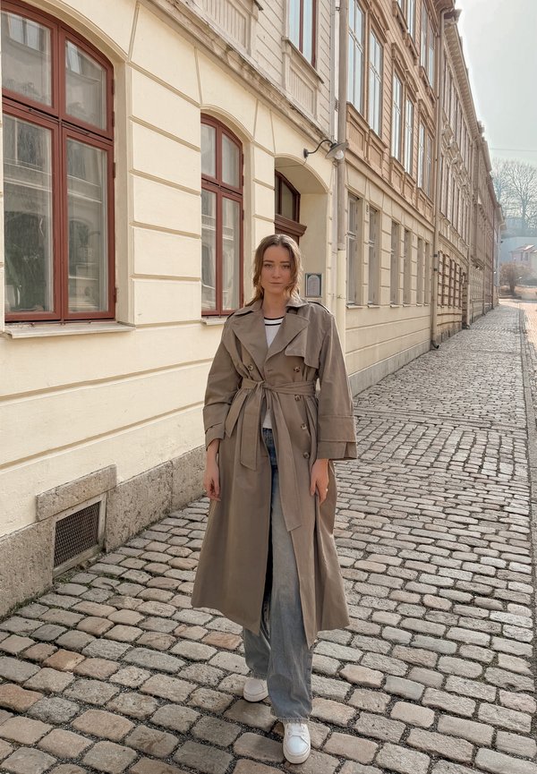Joven mujer con gabardina beige y jeans de pie en una calle adoquinada junto a un edificio color crema con ventanas arqueadas.