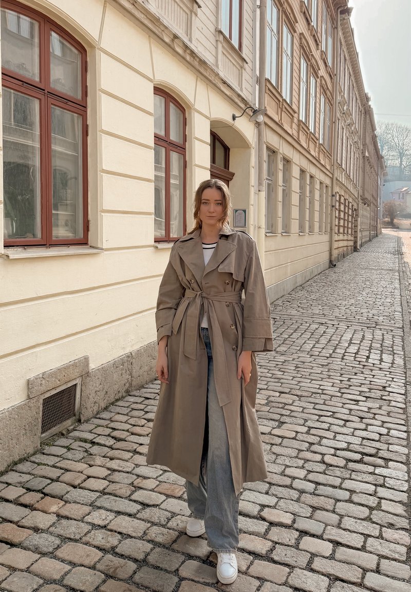 Giovane donna con trench beige e jeans, in piedi su una strada acciottolata accanto a un edificio color crema con finestre ad arco.