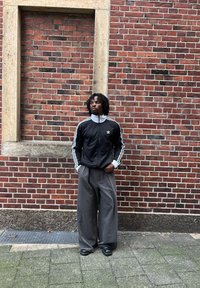 Černá bunda na zip s bílými pruhy, doplněná o šedé široké kalhoty. Pevné černé boty. Pozadí z cihlové zdi s dřevěným rámem.