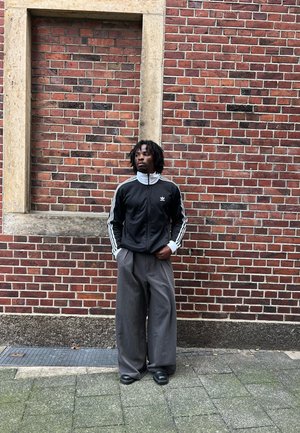 Chaqueta negra con cremallera y rayas blancas, combinada con pantalones de pierna ancha grises. Zapatos negros sólidos. Fondo de pared de ladrillo con un marco de madera.