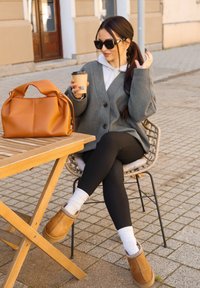 Smeđa kožna torbica, siva pletena kardigan, crne tajice, bež čizme do gležnja s dekorativnim rubom, bijele čarape, šalica kave u ruci, vanjsko okruženje.