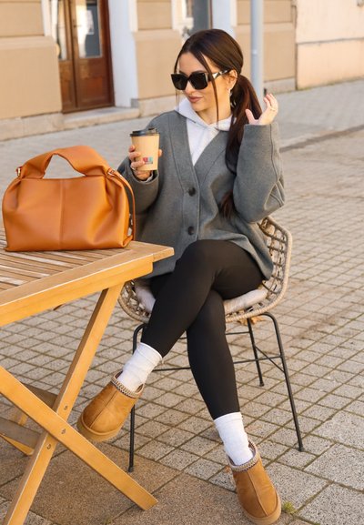 Bolso de mano de cuero marrón, cárdigan gris de punto, leggings negros, botines tawny con adornos decorativos, calcetines blancos, taza de café en la mano, entorno al aire libre.