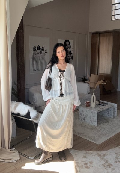 Joven mujer vestida con un conjunto blanco en capas y botas se encuentra en una sala de estar moderna con mesa de café de mármol y silla negra con piel sintética.