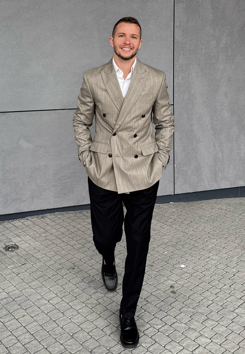Blazer bege riscado de dois botões sobre uma camisa branca, combinado com calças pretas e sapatos pretos brilhantes, a caminhar sobre um pavimento cinza.