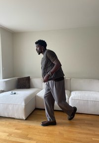 Mężczyzna w kamizelce, koszulce i spodniach, spacerujący wewnątrz obok białej kanapy modułowej z poduszką w panterkę i pilotami.