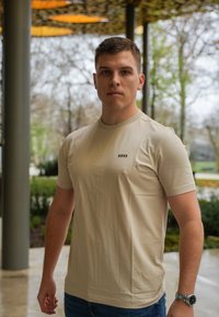 Beige t-shirt med kort ärm och rundad hals, med en liten "BOSS"-logotyp på bröstet. Bärs med blå jeans och en silverfärgad klocka.