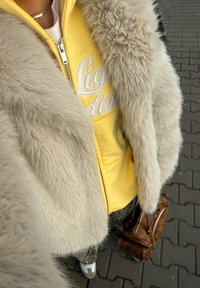 Casaco de peluche claro em bege sobre um hoodie amarelo com fecho éclair e letras brancas. Calças cinzentas com padrão e uma mala castanha. Tenis brancos.