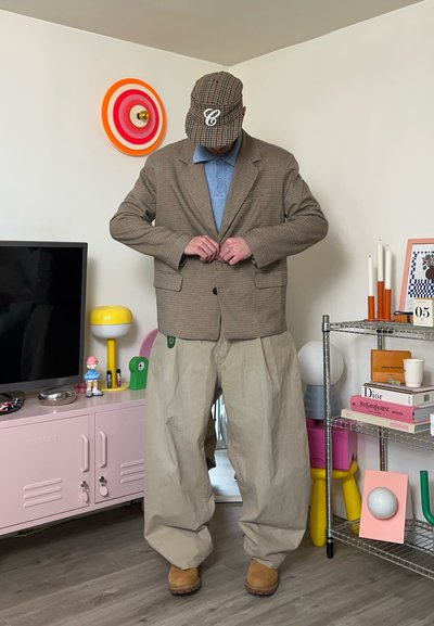 Hombre abrochándose un blazer marrón a cuadros, lleva una gorra de cuadros, un polo azul, pantalones anchos beige y botas tostadas en una habitación colorida con estanterías y decoración.