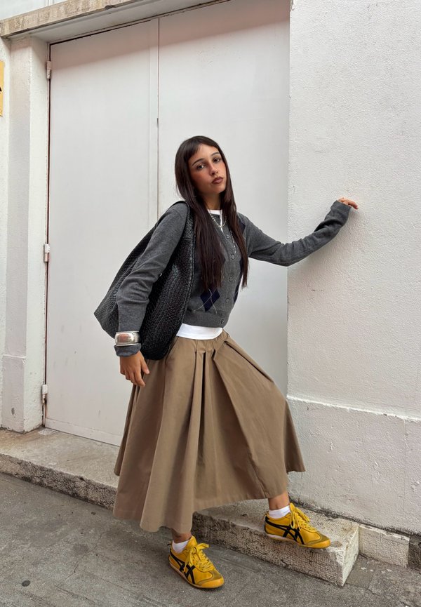 Una mujer posa junto a una puerta blanca. Lleva una chaqueta de punto gris, una falda larga de color beige y zapatillas deportivas amarillas, y lleva un bolso negro al hombro.