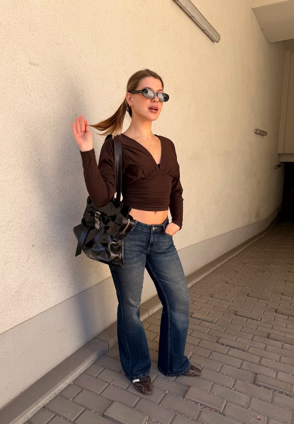Jeune femme portant un haut noir, un jean bleu, des lunettes de soleil foncées et portant un sac noir, se tient sur un chemin pavé à côté d'un mur beige.