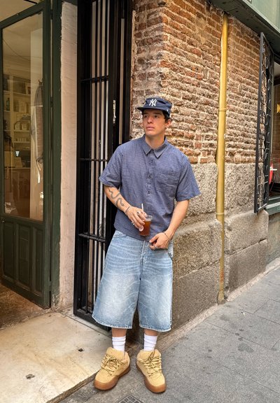 Camisa de cuadros azul de manga corta, pantalones cortos de mezclilla oversize claros, zapatillas color beige, calcetines blancos; sosteniendo una taza, de pie contra una pared de ladrillos.