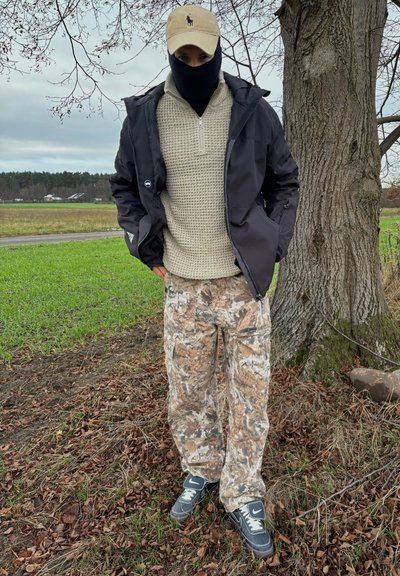 Chaqueta negra sobre un suéter de punto beige, pantalones de camuflaje y zapatillas negras. Gorra de un beige claro, cara cubierta con una balaclava negra.