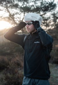 Veste polaire noire avec demi-fermeture éclair, gants d'extérieur, lunettes de soleil et un bonnet polaire blanc. Bretelles de sac à dos visibles. Fond naturel avec lumière douce.