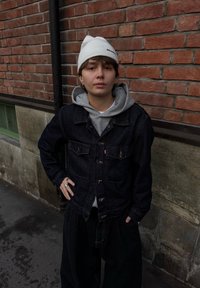 Temno modra denim jakna z dvema žepoma na prsih in svetlo siva jopa spodaj. Oblečena z ohlapnimi črnimi hlačami. V ozadju je opečnati zid.