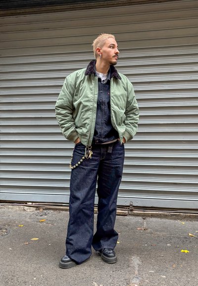 Cazadora bomber verde claro con cuello negro, peto vaquero azul y zapatos negros. Cadena plateada con cuentas sujetas al peto.