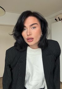 Femme aux cheveux noirs ondulés et maquillage léger, portant un pull blanc et un blazer noir à fines rayures, posant à l'intérieur contre un mur uni et des meubles.