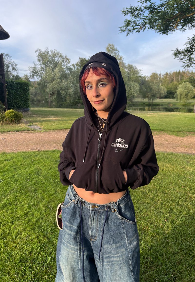 Jeune personne aux cheveux courts et rouges portant un sweat à capuche Nike Athletics noir et un jean ample, debout sur l’herbe près des arbres sous un ciel partiellement nuageux.