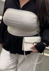 Γυναίκα που φοράει μαύρο μπλουζάκι, λευκό παντελόνι, λευκό τοπ τύπου ρούχου με ζώνη από πέρλες, κρατώντας μια μικρή λευκή τσάντα χειρός με λουρί αλυσίδας.