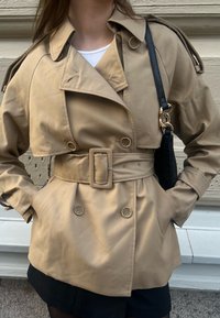 Beige trench coat med dubbelknäppt design, bälte i midjan, axelremmar och knäppta manschetter, tillsammans med en svart väska.