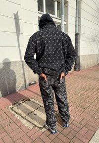 Casaco de ganga preto com logótipos repetidos, combinado com calças pretas padrões com texto e gráficos. Ténis cinzentos com realces azuis.