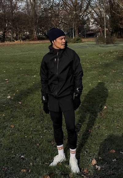Chaqueta negra impermeable con capucha, pantalones cortos negros ajustados, guantes y zapatillas deportivas blancas. La escena está ambientada en un parque con césped y árboles.
