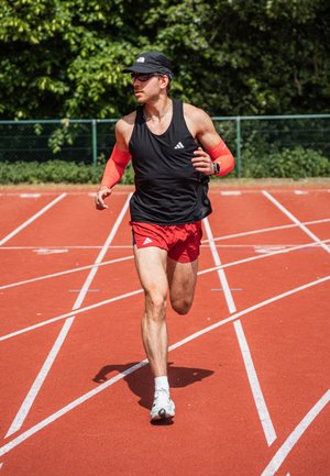 Atleta in top senza maniche nero e shorts rossi, con maniche rosse e cappellino, che corre su una pista rossa con segnaletica bianca.