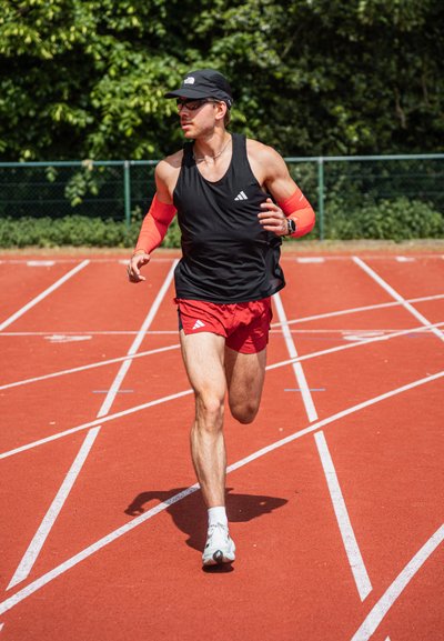 Atleta con camiseta sin mangas negra y pantalones cortos rojos, que lleva mangas rojas y una gorra, corriendo en una pista roja con marcas blancas en los carriles.