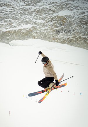 Esquiador realizando un salto con esquís multicolores de superficie texturizada, llevando casco y guantes negros, sobre un fondo nevado.