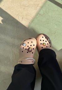 Beige Crocs med dekorative charms som inkluderer en fargerik blomst, et tegneserieansikt, et kraftsymbol og et perlearmbånd på venstre stropp.
