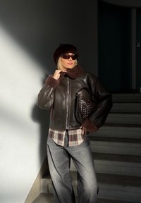 Casaco de pele castanha com gola de pelo, camisa aos quadrados por baixo, calças largas cinzentas e uma mala entrelaçada castanha escura, numa escadaria bem iluminada.