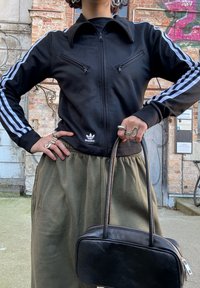 Fekete Adidas szabadidőfelső fehér csíkokkal az ujjakon, cipzáros zsebekkel, mellette olívazöld szoknya és egy kis fekete kézitáska.