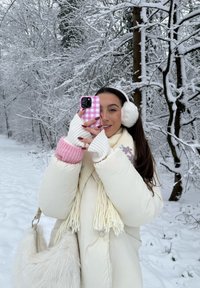 Casaco branco fofo, mangas de malha rosas e brancas, cachecol creme com franjas e uma mala de pelo branco. Segurando uma capa de telemóvel xadrez rosa numa floresta coberta de neve.